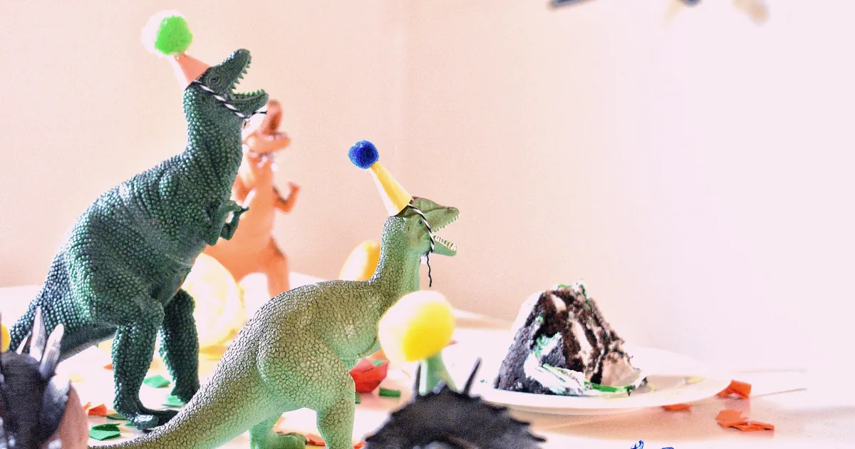 Toy dinosaurs in paper party hats gathered on a confetti-strewn table beside a slice of chocolate cake.