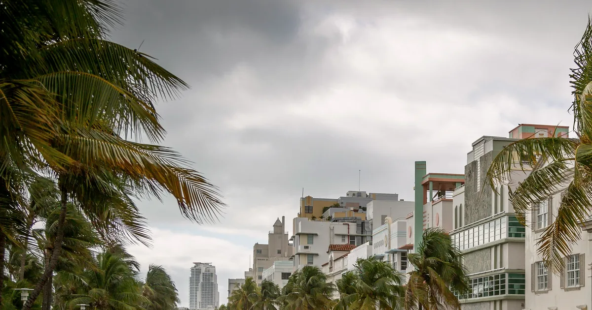 Storm clouds over South Beach, palm trees bending in the wind beside pastel Art Deco buildings.